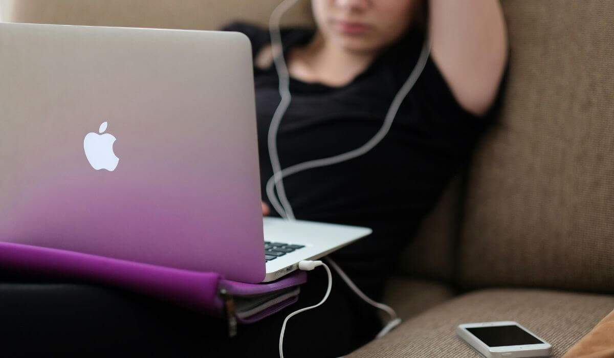 Teen sitting on a couch using a laptop and headphones, illustrating the impact of social media and gadget usage on the mental health of teens.