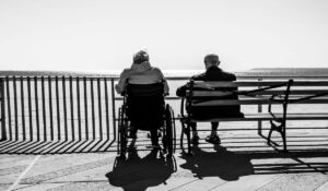 An elderly couple sitting by the seaside, with one in a wheelchair, symbolizing the emotional challenges caregivers face, potentially leading to compassion fatigue.