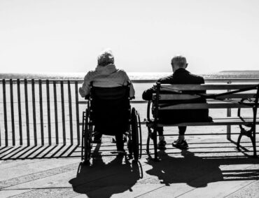 An elderly couple sitting by the seaside, with one in a wheelchair, symbolizing the emotional challenges caregivers face, potentially leading to compassion fatigue.