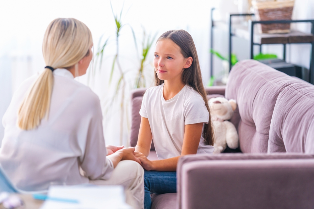 A woman and a girl are sitting on a purple sofa in a bright room, engaging in what seems like parent-child counseling