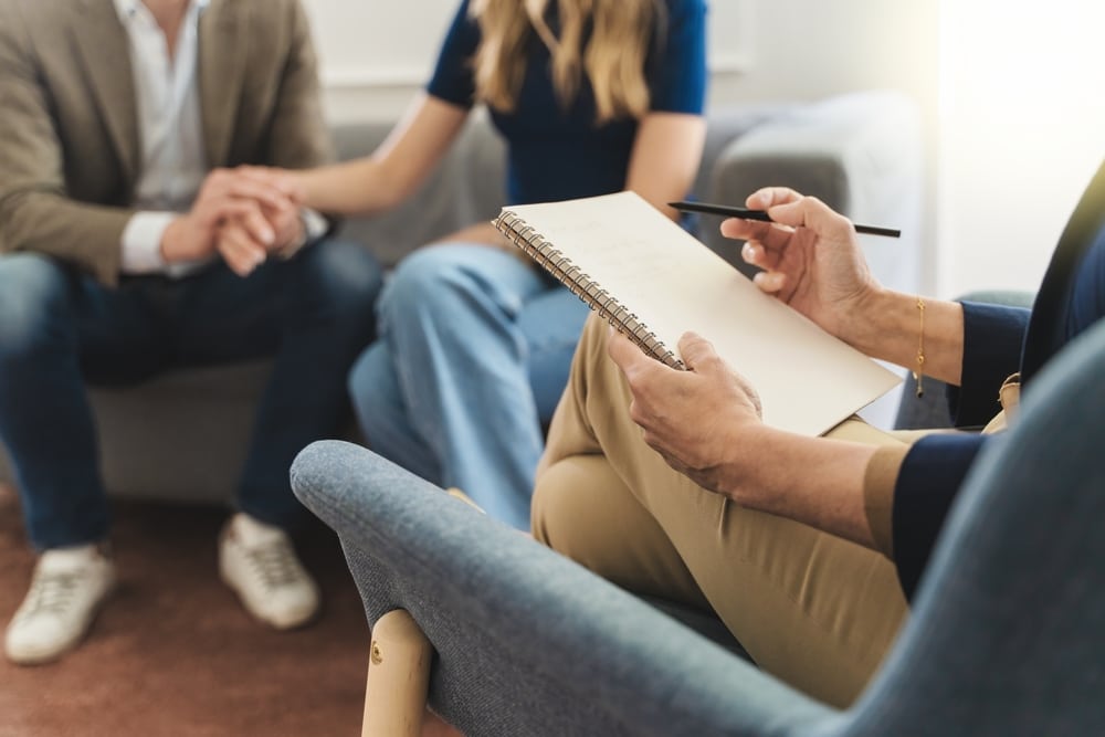 A woman sits in a chair, writing notes on a notepad during a couples counseling session