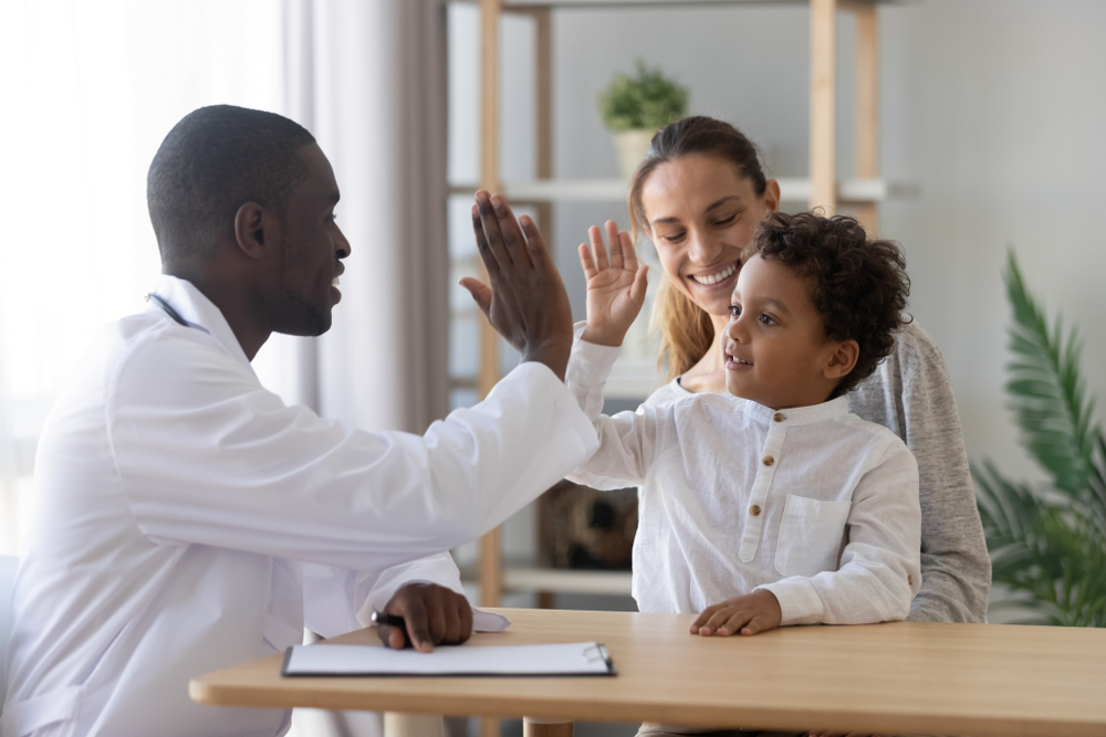 A doctor in a white coat high-fives a young boy sitting with his mother at a table, showcasing the warmth often found in parent-child counseling sessions