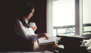 A woman drinking coffee and reading by the window, symbolizing social isolation and chronic loneliness.