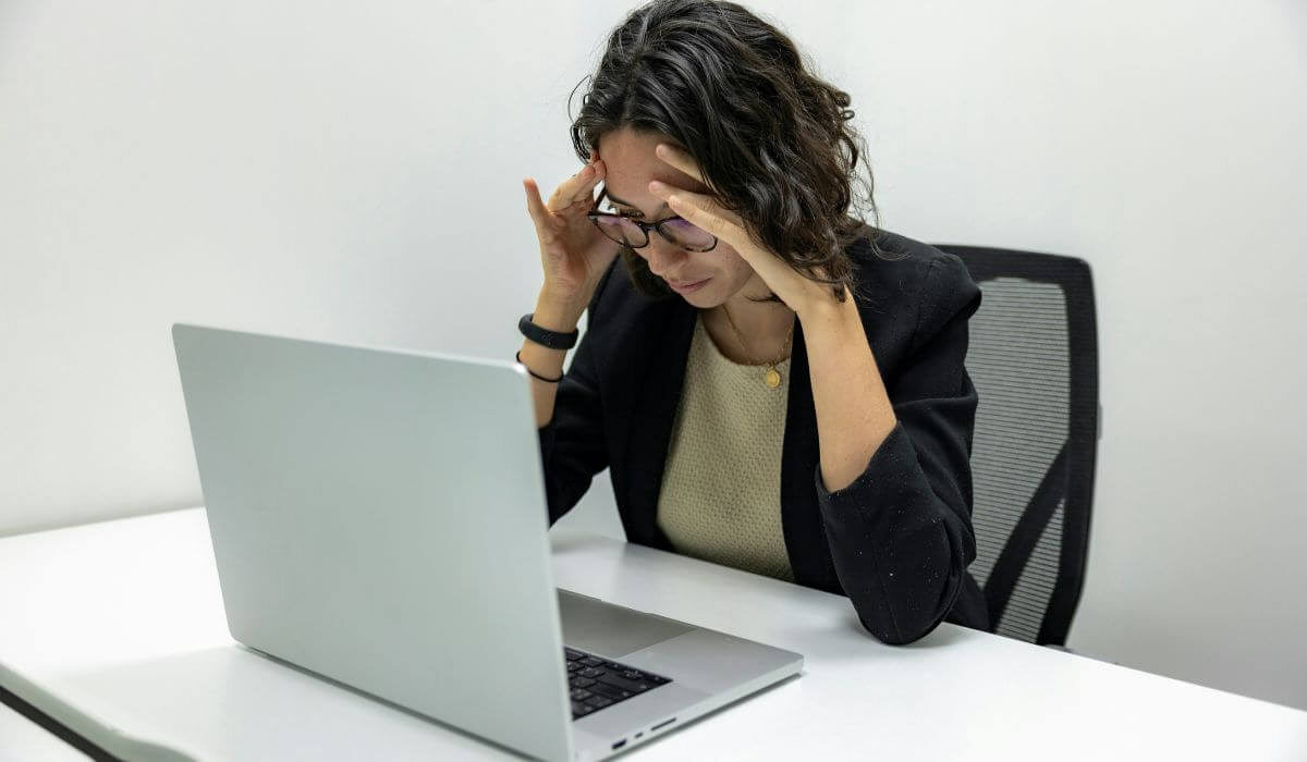 A woman sitting at a desk, holding her forehead in frustration, symbolizing mental health challenges and the need for burnout therapy.