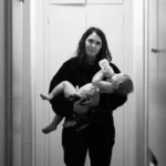 Exhausted mother bottle-feeding baby in hallway, wondering if it’s time to talk to a therapist.