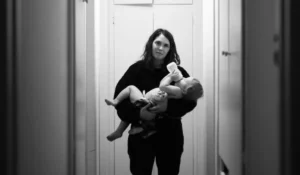 Exhausted mother bottle-feeding baby in hallway, wondering if it’s time to talk to a therapist.