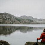 A person in a red coat sits alone by a quiet lake, reflecting near the mountains – representing emotional calm and trauma therapy.