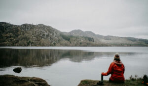 A person in a red coat sits alone by a quiet lake, reflecting near the mountains – representing emotional calm and trauma therapy.