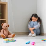Young girl showing signs of childhood anxiety while sitting alone in a quiet, toy-filled playroom corner.
