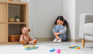 Young girl showing signs of childhood anxiety while sitting alone in a quiet, toy-filled playroom corner.