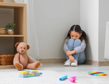 Young girl showing signs of childhood anxiety while sitting alone in a quiet, toy-filled playroom corner.