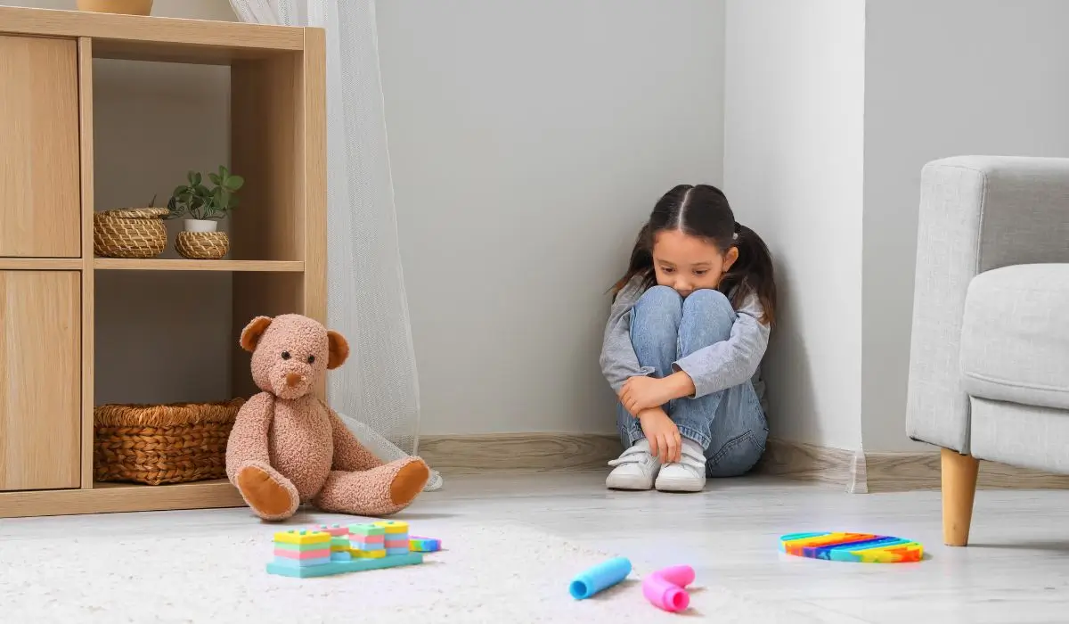 Young girl showing signs of childhood anxiety while sitting alone in a quiet, toy-filled playroom corner.