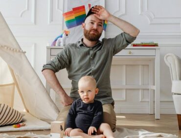 First-time father seated indoors, looking fatigued while caring for his baby—capturing the emotional weight of early fatherhood.