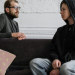 Teen wearing hoodie in counseling session with adult therapist on a couch, representing teen therapy Glen Ellyn and safe emotional check-ins.