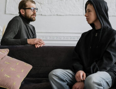 Teen wearing hoodie in counseling session with adult therapist on a couch, representing teen therapy Glen Ellyn and safe emotional check-ins.