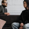 Teen wearing hoodie in counseling session with adult therapist on a couch, representing teen therapy Glen Ellyn and safe emotional check-ins.